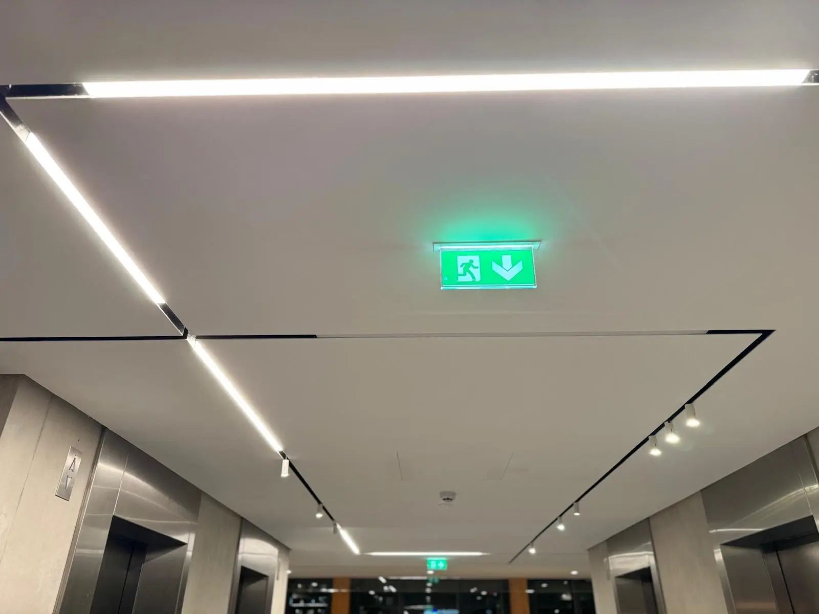 Hall d'ascenseurs : plafond blanc, joints creux sombres et réglettes lumineuses