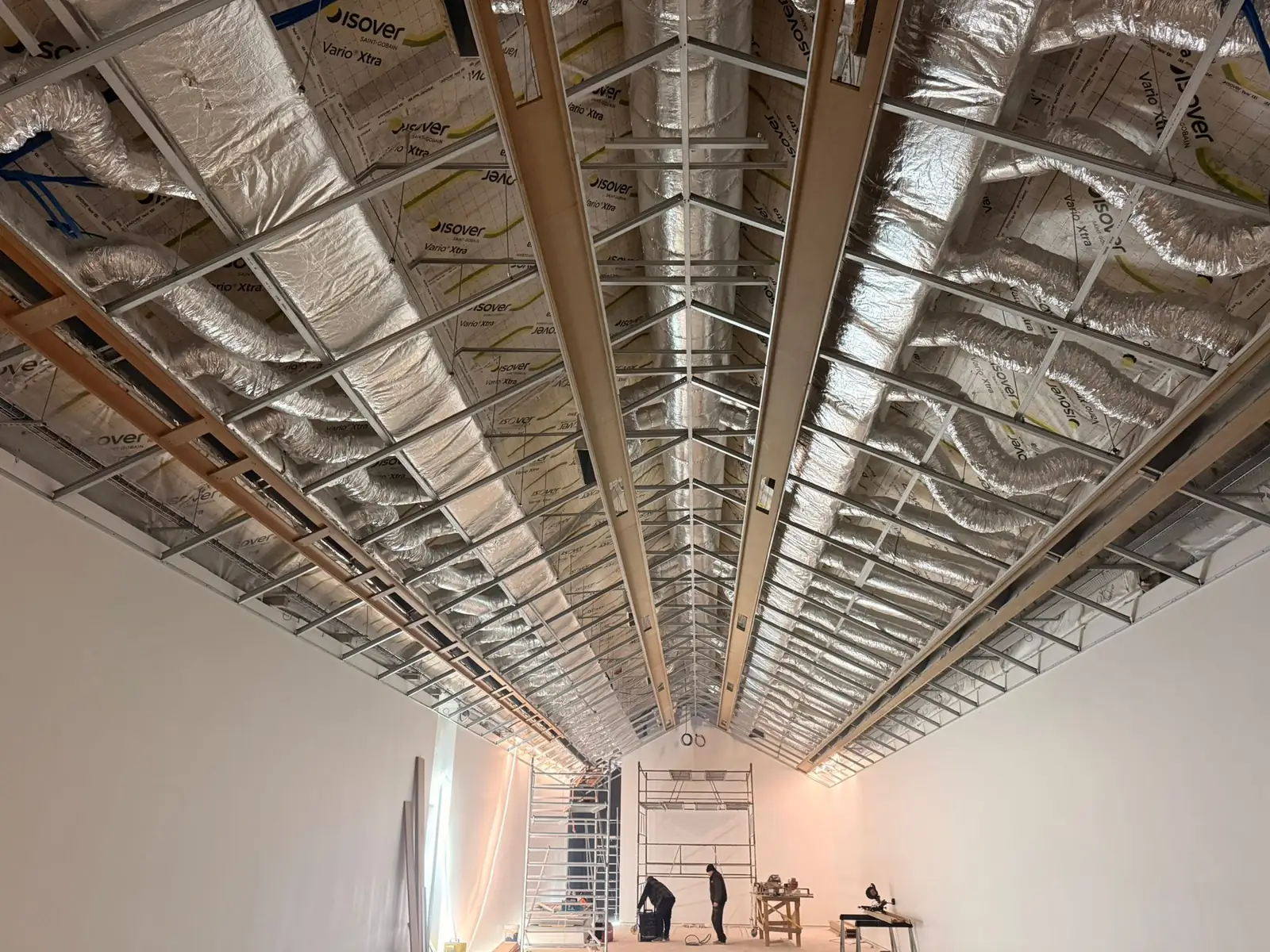 Grande salle du musée en chantier : ossature métallique du plafond sur toute la longueur, isolant Isover posé entre les profilés, gaines de ventilation souples, deux compagnons à l'échafaudage au fond