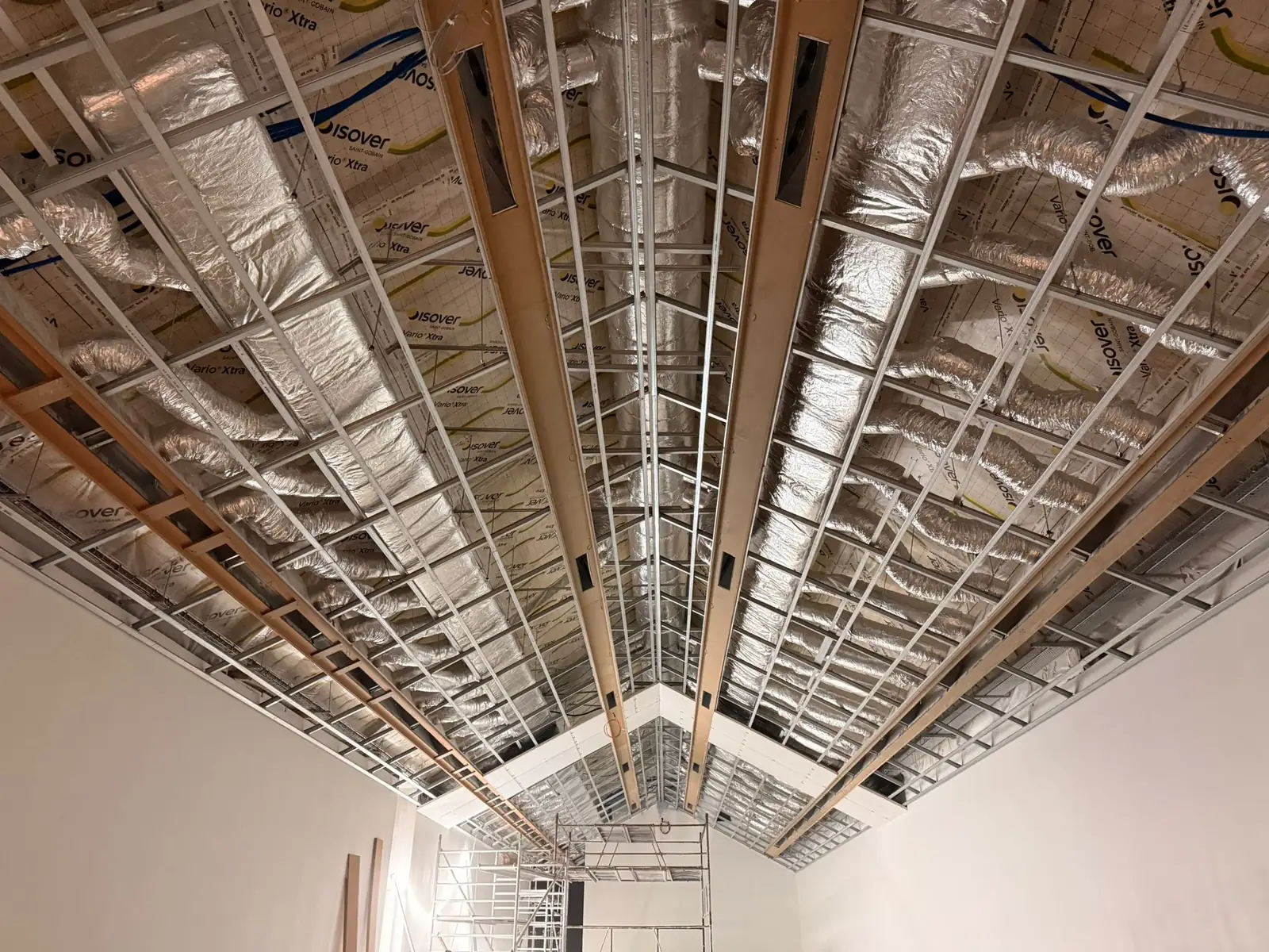 Contre-plongée sur l'ossature du plafond : profilés métalliques alignés, isolant en pare-vapeur réfléchissant, gaines de ventilation et poutres bois filant vers le faîtage
