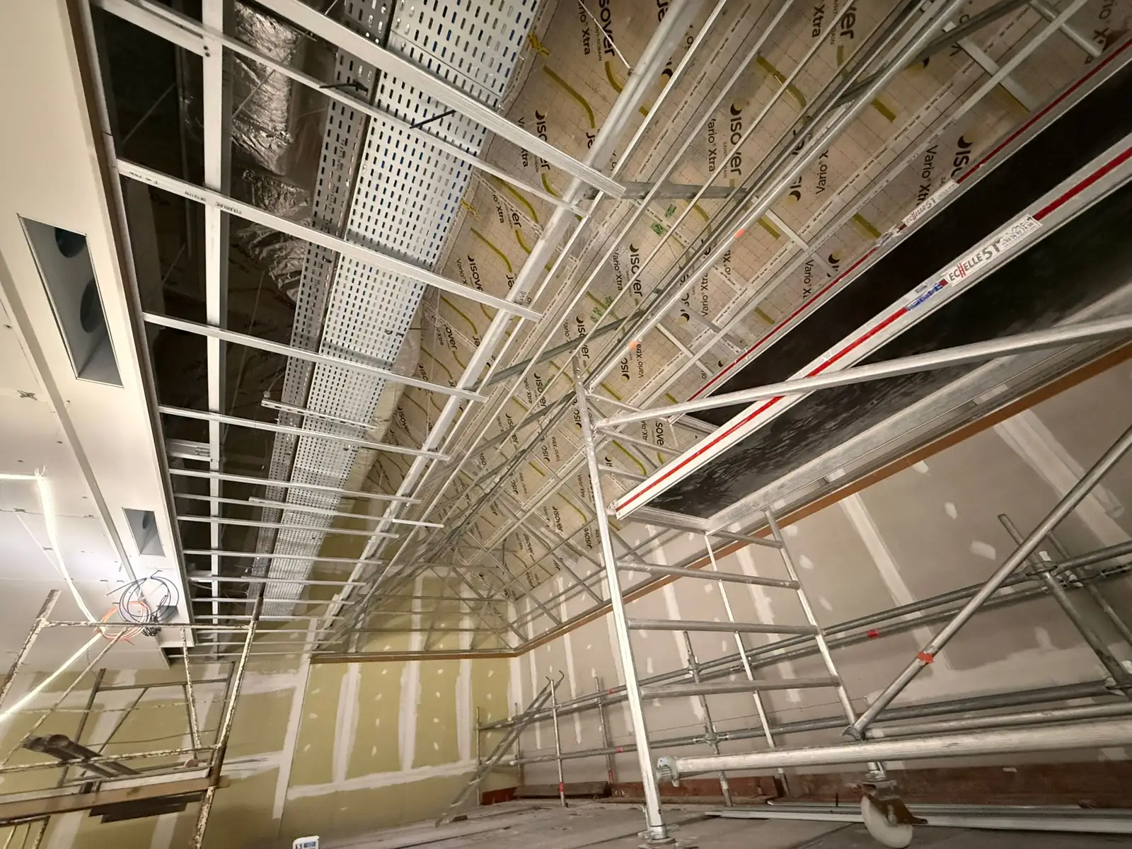 Ossature de plafond en cours de pose le long d'un mur, échafaudage roulant en place, chemins de câbles et retombée déjà plaquée à gauche