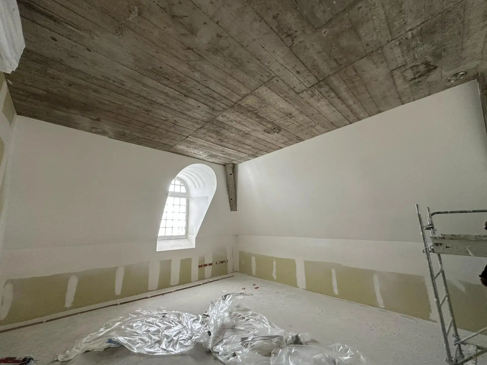 Pièce des combles en fin de chantier : dalle de béton coffrée en planches laissée apparente, murs plaqués et enduits, lucarne à ébrasement cintré, bas de cloison encore en plaque avant enduit, échafaudage roulant à droite