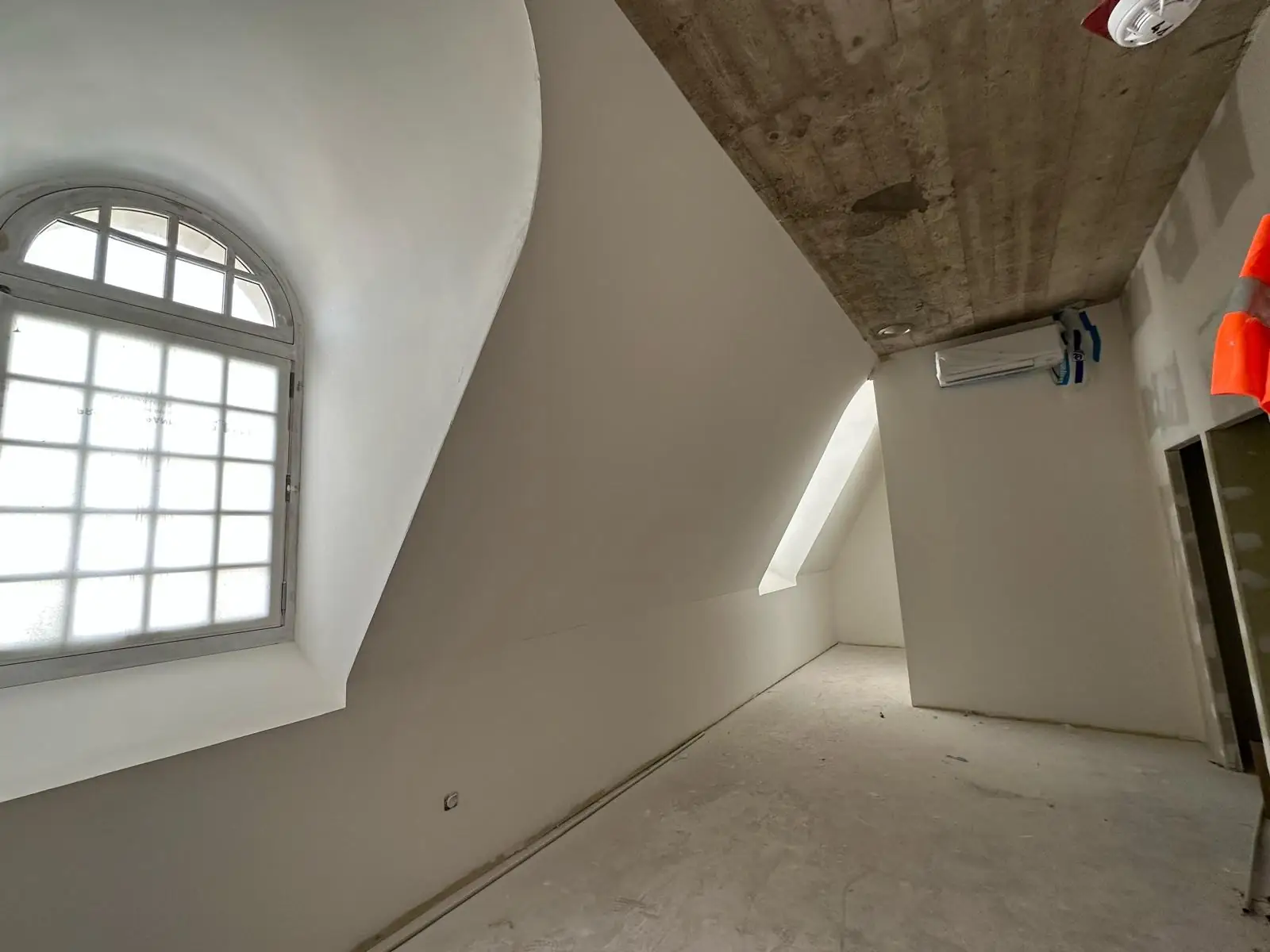 Ébrasement de lucarne cintré en plâtre lissé, courbe continue du tableau jusqu'au linteau ; au fond, enfilade des combles sous la dalle de béton coffrée conservée apparente