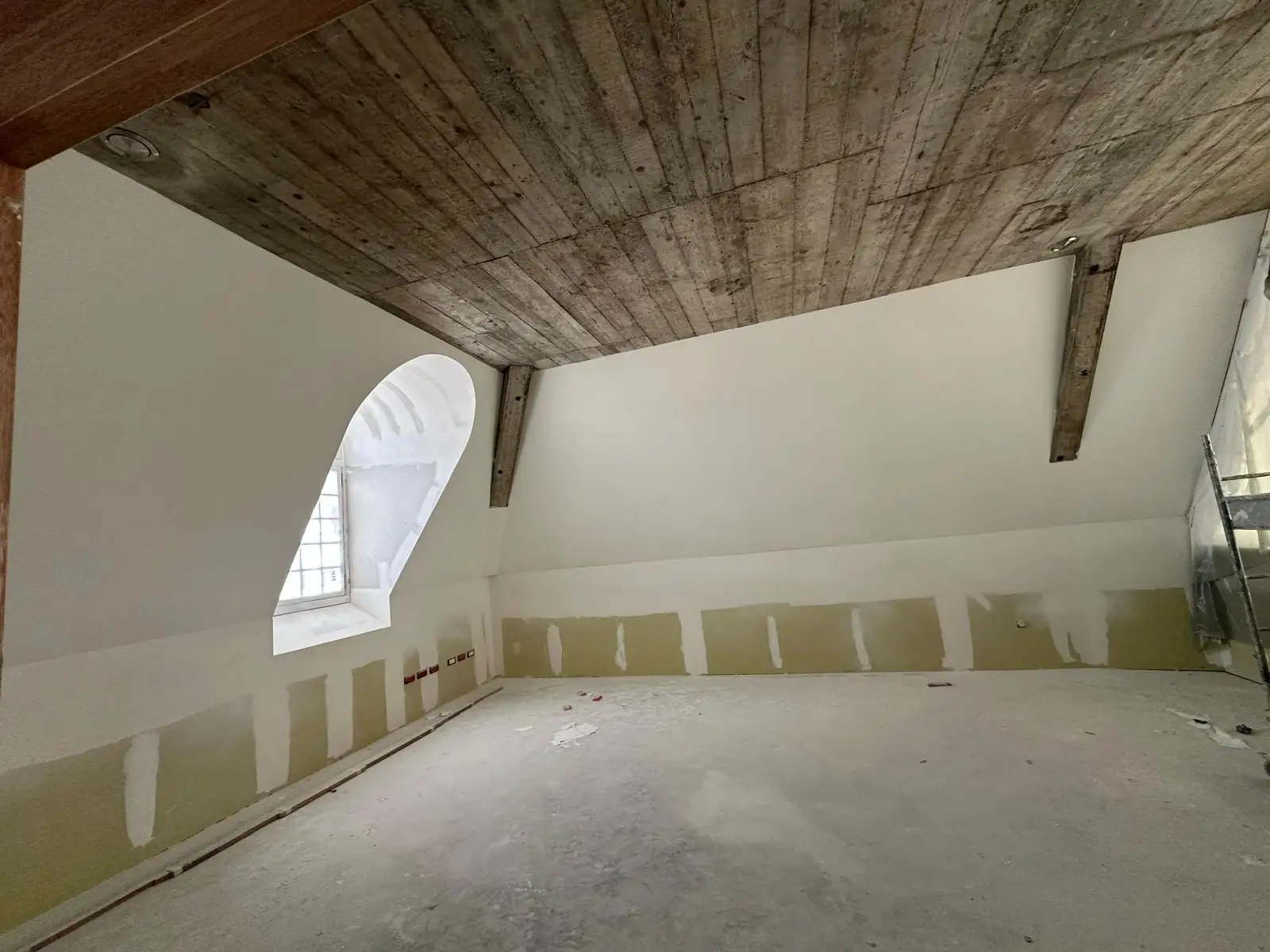 Pièce des combles sous dalle de béton coffrée et poutres apparentes, lucarne cintrée ouverte sur le toit, bande basse des cloisons encore en plaque, joints enduits par passes verticales
