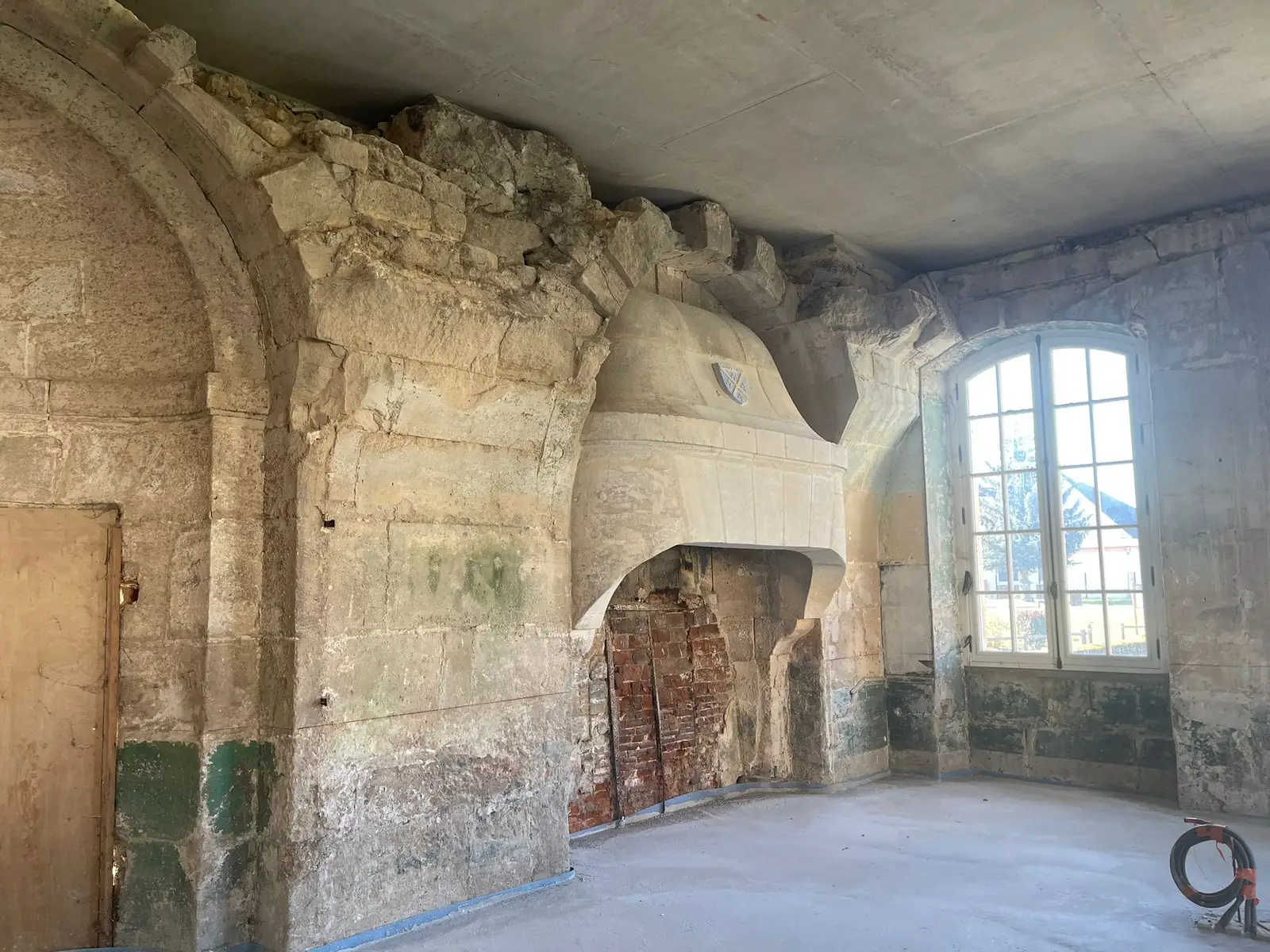 Salle avant travaux : cheminée monumentale en pierre sous un arc de décharge appareillé, murs décapés jusqu'à la pierre, fenêtre à petits bois